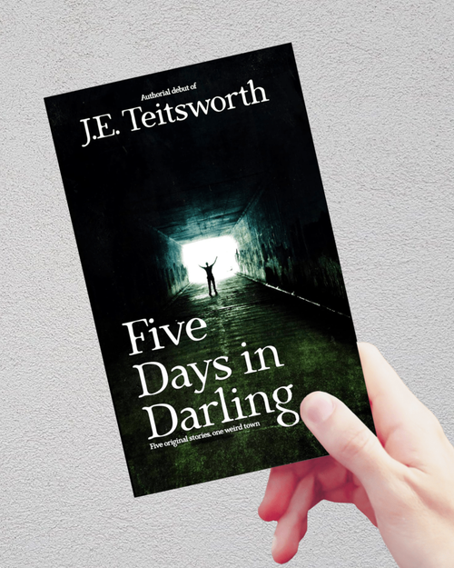 A hand holding a paperback book titled 'Five Days in Darling' by J.E. Teitsworth against a light gray textured wall.