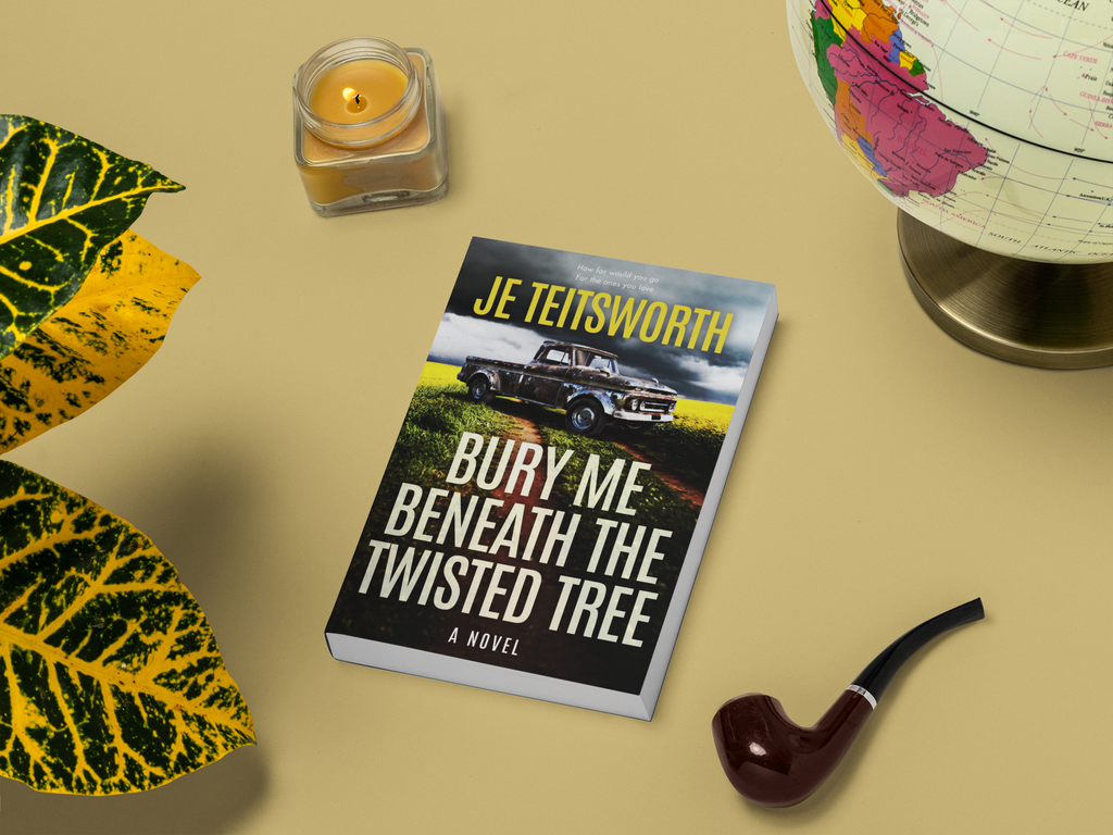 A book titled 'Bury Me Beneath the Twisted Tree' by JE Teitsworth is displayed on a beige surface with decorative props, including a lit candle, a globe, a pipe, and green and yellow leaves.