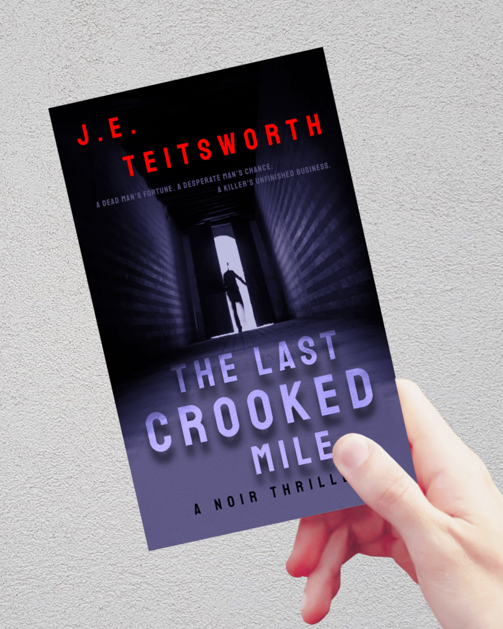 A hand holding a book titled 'The Last Crooked Mile' by J.E. Teitsworth against a textured light gray wall.